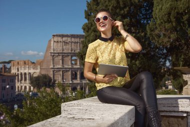 arkadan görünüşü parapetin üzerinde oturmuş ve ses kılavuzu Tablet Pc yakınlarında Kolezyum, Roma, İtalya, dinlerken turist kadın 