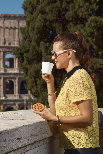 smiling young woman in yellow blouse drinking coffee and holding Italian mini crostata near Colosseum , Rome, Italy