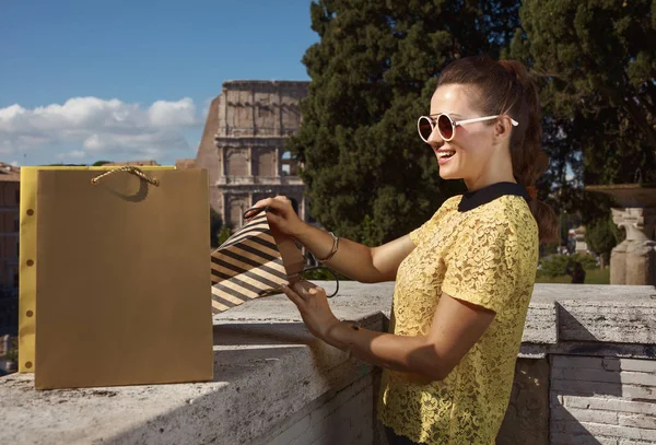 smiling modern woman in yellow blouse looking in shopping bag with Colosseum on background, Rome, Italy