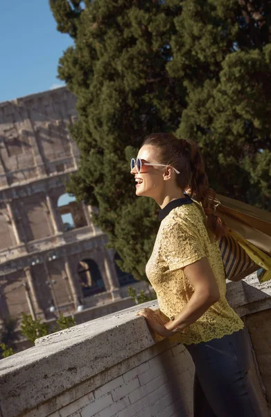 happy modern woman in yellow blouse with shopping bags looking into distance and leaning on parapet near Colosseum , Rome, Italy