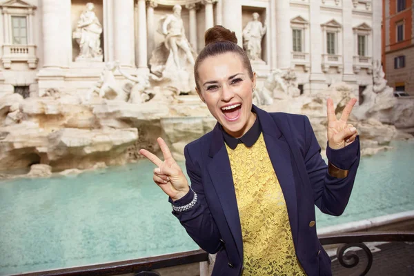 happy modern woman in yellow blouse and blue jacket showing victory gesture near Trevi Fountain, Rome, Italy