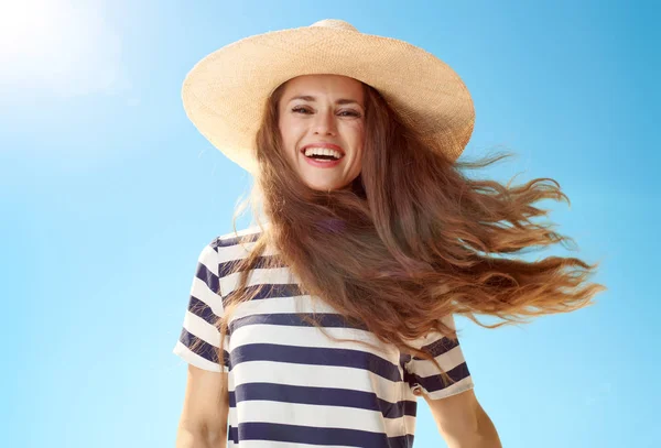 Portrait of smiling trendy woman in straw hat with fluttering hair against blue sky