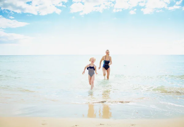 海岸で海から実行されている水着はモダンな母と子の笑顔の完全な長さの肖像画 — ストック写真