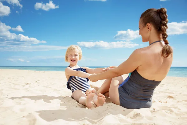 微笑的现代母亲和孩子在泳衣上玩耍 而坐在海边的沙滩上 — 图库照片