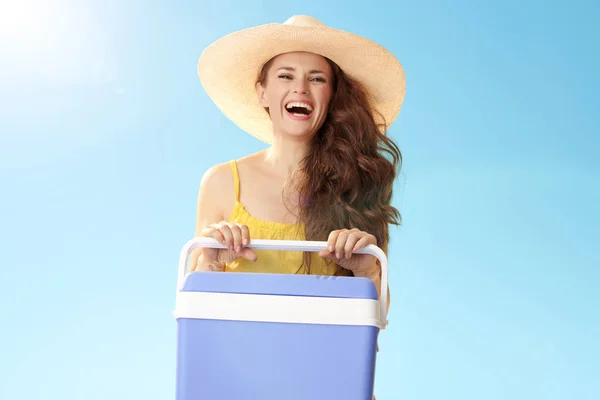 happy active woman in straw hat with plastic cooler box laughing against blue sky