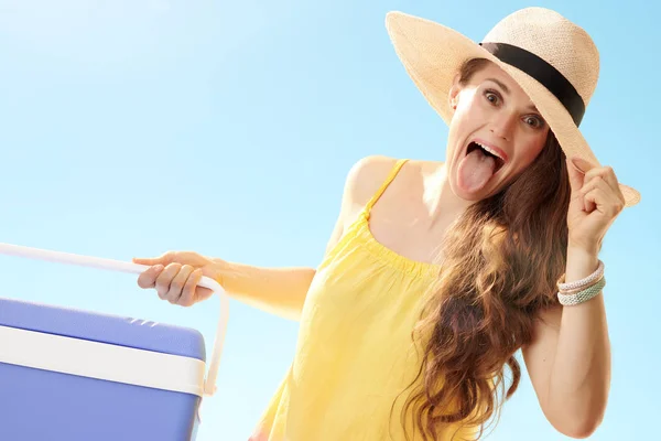 愉快的妇女在草帽与蓝色塑料凉快的箱子有乐趣时间和显示舌头反对蓝色天空 — 图库照片