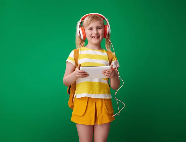 smiling pupil with backpack holding tablet PC and listening to music on green background