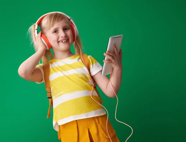 微笑的女孩与背包举行平板电脑和听音乐在耳机上的绿色背景 — 图库照片