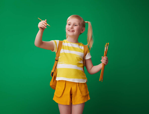 smiling school girl with backpack drawing with brush in the air on green background 