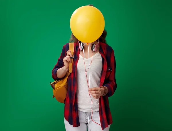 バックパックと緑の背景の顔の前に黄色のバルーンを保持しているヘッドフォンと赤シャツのモダンな学生女性を笑顔 — ストック写真