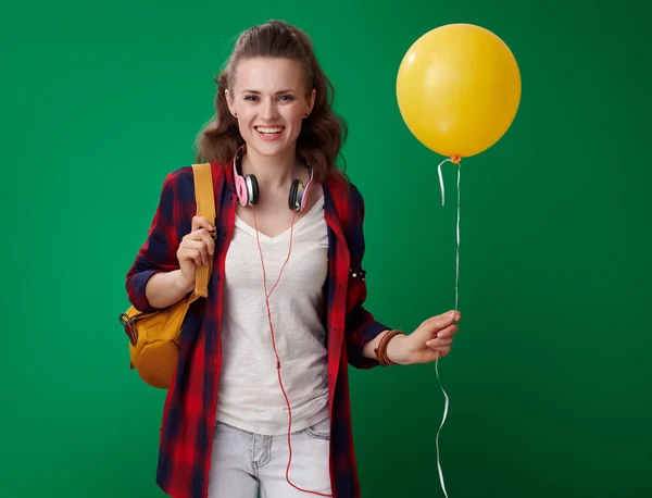 バックパックと黄色のバルーンを押しながら緑の背景にカメラ目線のヘッドフォンと赤シャツ幸せな若い学生女性 — ストック写真