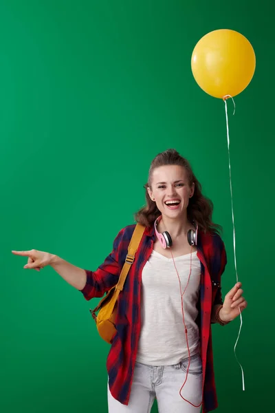 バックパックとヘッドフォン黄色のバルーンを押しながら脇を指している緑の背景に赤のシャツで幸せなモダンな学生女性 — ストック写真