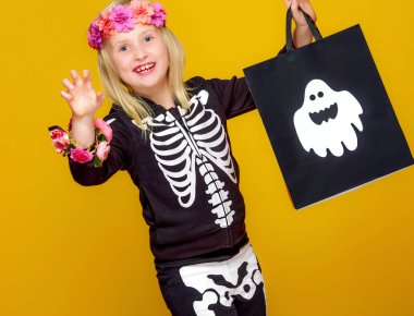 smiling girl in halloween skeleton costume showing shopping bag and looking at camera while frightening on yellow background clipart