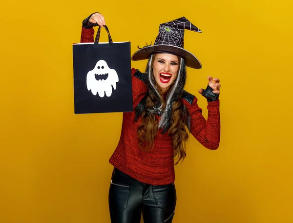 ハロウィン魔女の買い物袋を示し 黄色の背景に恐ろしいしながらカメラ目線で現代女性の笑みを浮かべてください — ストック写真