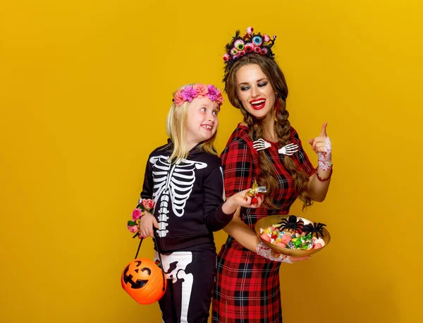 背景が黄色のキャンディーとジャック ランタンのプレートを保持しているメキシコ スタイルのハロウィーンの衣装は現代の母と子の笑顔 — ストック写真