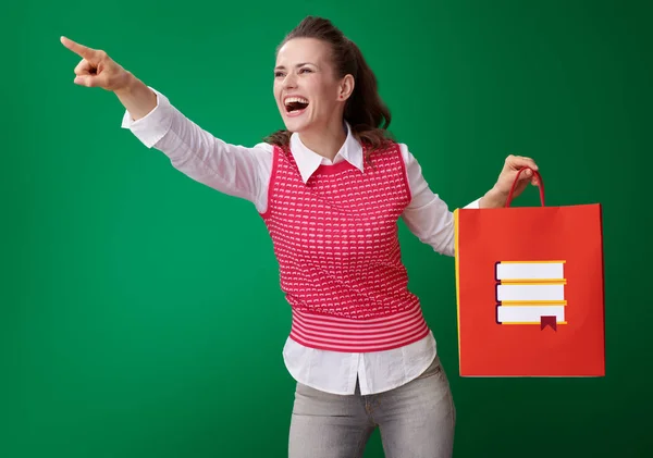 本ショッピング バッグを押しながら脇を指している緑の背景に赤のチョッキで幸せな若い学生女性 — ストック写真