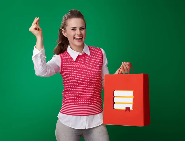 赤いチョッキ本ショッピング バッグを押しながら緑の背景に対して指をスナップで幸せなモダンな学生女性 — ストック写真