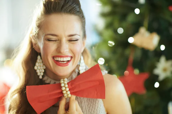 微笑着时尚的家庭主妇与闭着眼睛在圣诞树附近拿着红色晚餐餐巾以领结的形式 — 图库照片