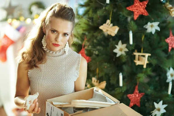 不愉快的时髦的女人附近的圣诞树拉出破碎的菜从包裹和看着相机 — 图库照片