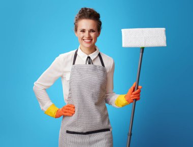 Portrait of happy young cleaning lady in apron with mop against blue background. Professional woman cleaner will provide a simple and hassle-free service clipart