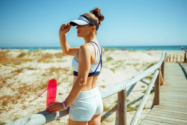 healthy sports woman in sport clothes with red bottle of water looking into the distance on the seashore. Outdoor fresh air excessing fore more energy. woman holding visor of cap