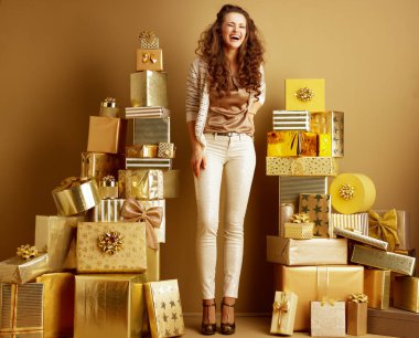 Full length portrait of happy stylish fashion-monger in beige clothes standing laughing with closed eyes and standing among pile of golden gifts in front of plain wall  clipart