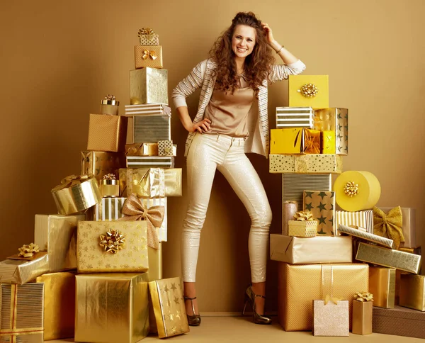 全长的肖像微笑的现代妇女在金色米色的衣服站在两堆金色的礼物在一个普通的墙壁前 在一个地方的效率购物的概念 生日插图 — 图库照片