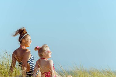 smiling young mother and daughter in swimwear on the seashore looking aside. quiet vacation heaven. protect your hair from sun, heat, and humidity before heading to the beach. blond hair child. clipart