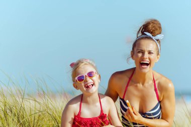 gülümseyen modern anne ve çocuk beachwear içinde ruj spf ile sahil üzerinde kullanın. Dudak kremi Spf güneşten korunma kuru dudaklar mücadele ile. Mavi gökyüzü. kalabalık barış için çok az. sessiz tatil cenneti.