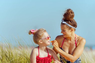 Modern anne ve kızı ruj spf ile okyanus kıyısında uygulama Mayo içinde gülümseyen. Dudak kremi Spf güneşten korunma kuru dudaklar mücadele ile. Güneş saç korumalı. kalabalık barış için en az.