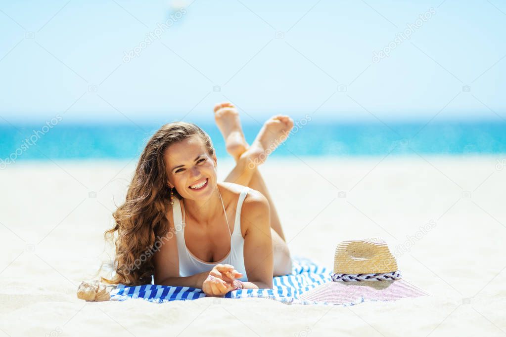 Portrait of smiling modern woman in white swimwear on the seacoast lying on a striped towel. straw hat laying on sand. Sun protected hair. Sunny summer midday.