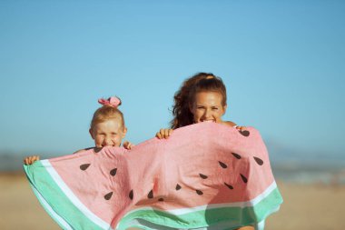 mutlu genç anne ve kızı mayo eğleniyor akşam sahilde komik karpuz havlu tutarken zaman. Güneş saç korumalı. Güneşli yaz akşam. Mavi gökyüzü.