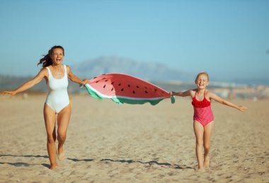 Modern anne ve çocuk mayo sahil akşam holding komik karpuz havlu üzerinde gülümseyen ve çalışıyor. kalabalık barış için çok az. Sarı saçlı çocuk çiçekli kırmızı noktalı mayo