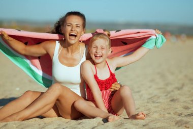 Modern anne ve çocuk beachwear sahil üzerinde kum üzerinde otururken komik karpuz havlu tutan akşam gülümseyerek. Plaj zaman - aktif ve dış yolu olmak mükemmel çocuklar ile.