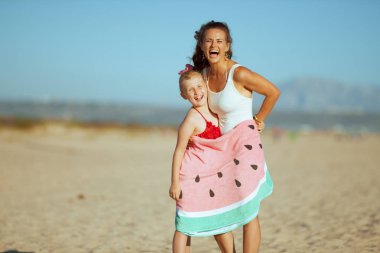 Modern anne ve kızı beachwear okyanus kıyısında içinde komik karpuz havlu sarılı akşam gülümseyerek. Güneş saç korumalı. Tüm aile için eğlenceli plaj-dostu etkinlikleri.