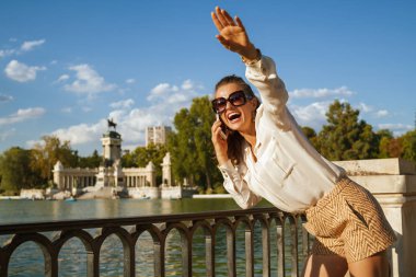 arkadaşlarıyla takılmak için bir smartphone kullanarak ve birine sallayarak zarif Gezgin kadın beyaz bluz ve El Retiro Park Madrid, İspanya, şort ya da yerel genç parti kadın gülümseyerek karşılayan.