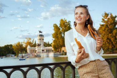 happy stylish traveler woman in white blouse and shorts at El Retiro Park in Madrid, Spain eating traditional Spain churro. churros - classic Spanish sweet treat. caucasian woman. blue sky. solo  clipart