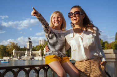 son moda anne ve çocuk gezginler, Parque del Buen Retiro Madrid, İspanya, bir şeye işaret içinde gülümseyen. Madrid İspanya güneşli başkentidir. Madrid Aile gezisi. Mikro Seyahat sahip. Mavi gökyüzü.