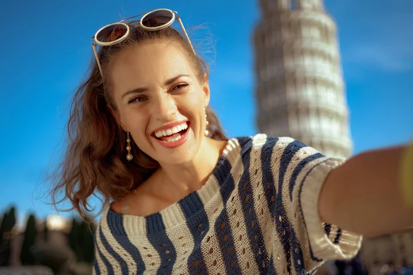 happy trendy woman in striped blouse in the front of leaning tower in Pisa, Italy taking selfie. classic example of overtourism. european woman brunette ponytail hairstyle 30 something years old.