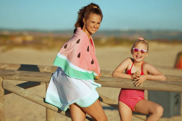 Mutlu genç anne ve çocuk akşam sahil üzerinde mayo portresi. Anne ve kızı ahşap bir çit yakınındaki. Saç güneş korumalı.