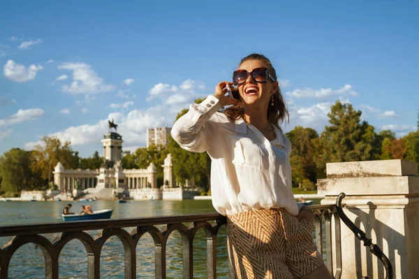 smiling modern woman in white blouse and shorts at Parque del Buen Retiro in Madrid, Spain talking on a cell phone. big fashionable sunglasses. woman talking to family or loved one & share experience.