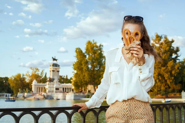 cheerful young traveler woman in white blouse and shorts at El Retiro Park in Madrid, Spain looking through traditional Spain churro. you cant leave Madrid without trying churros. blue sky.