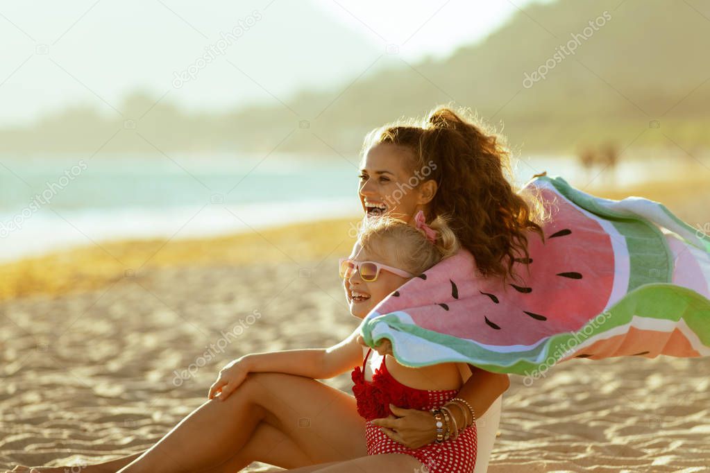 happy modern mother and child in beachwear holding watermelon towel sitting on the seashore in the evening having fun time. minimal to no crowd peace. blond hair child wearing sunglasses.