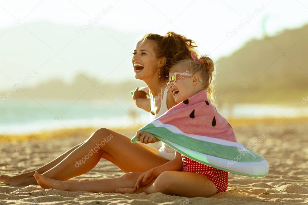 happy modern mother and child in swimwear on the seashore in the evening having fun time wrapped in watermelon towel. minimal to no crowd peace. Fun beach-friendly activities for the whole family.