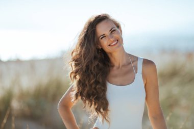 Deniz kıyısı üzerinde beyaz mayo mutlu modern kadın portresi. yeşil çim ve hiçbir insan ile vahşi plaj. Mavi gökyüzü. kalabalık barış için çok az. Güneş saç korumalı. Avrupa kadın.