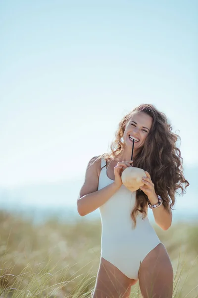 ココナッツから飲んでビーチで白の水着で幸せな若い女 ココナッツの栄養は 免疫システムに最適です ビーチの職業 緑の草とは人々 と野生のビーチ 青い空 — ストック写真