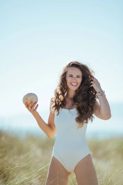 happy modern woman in white swimwear on the ocean shore holding coconut. Coconut oil as a natural way to help your hair grow longer, thicker, and faster. blue sky. quiet vacation heaven.