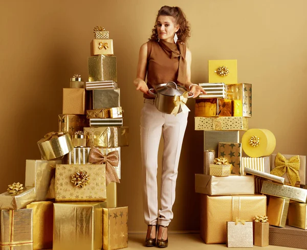 Full length portrait of unhappy young shopper woman in gold beige pants and brown blouse with a stew pan with golden bow as a received bad gift among 2 piles of golden gifts. bad present concept.