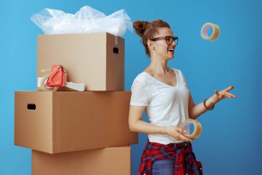 smiling modern woman in white t-shirt near cardboard box throwing up adhesive tapes isolated on blue clipart