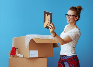 smiling modern woman in white t-shirt near cardboard box looking at the photo frame against blue background clipart